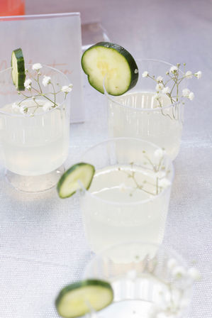 Refreshing cucumber lemonade in clear plastic cups, each garnished with a cucumber slice and delicate white baby's-breath flowers on a light textured tablecloth — perfect summer party or brunch drink.