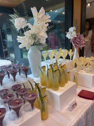 Reception cocktail table at an indoor event with purple dessert martinis topped with gold flakes, tall pineapple‑garnished tropical drinks, lemon martinis with lemon wheels arranged on white risers and accented by white and blush floral arrangements in ceramic vases.