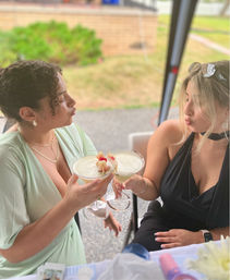 Two friends toasting with frothy, fruit‑garnished cocktails at an outdoor patio table, playful puckered faces, one in a mint dress and one in a black halter, blurred lawn in the background.