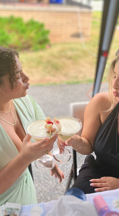 Two friends toasting with frothy, fruit‑garnished cocktails at an outdoor patio table, playful puckered faces, one in a mint dress and one in a black halter, blurred lawn in the background.