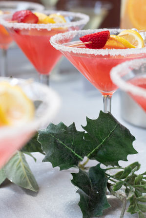Sugar-rimmed pink cocktails in martini glasses garnished with strawberries and lemon slices beside green decorative leaves on a party table.