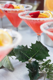 Sugar-rimmed pink cocktails in martini glasses garnished with strawberries and lemon slices beside green decorative leaves on a party table.