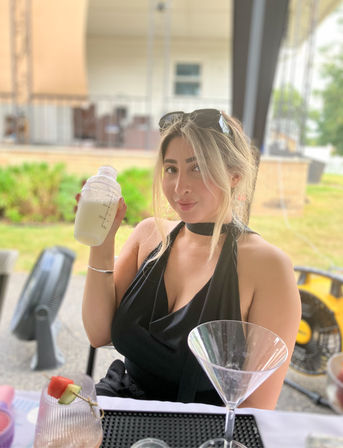 Woman in a black halter top holding a cocktail shaker with a creamy drink at an outdoor patio bar, martini glass and fruit skewer on the table, sunglasses perched on her head.