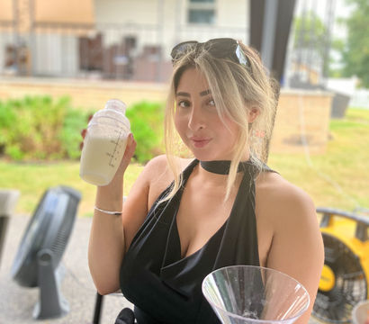 Woman in a black halter top holding a cocktail shaker with a creamy drink at an outdoor patio bar, martini glass and fruit skewer on the table, sunglasses perched on her head.