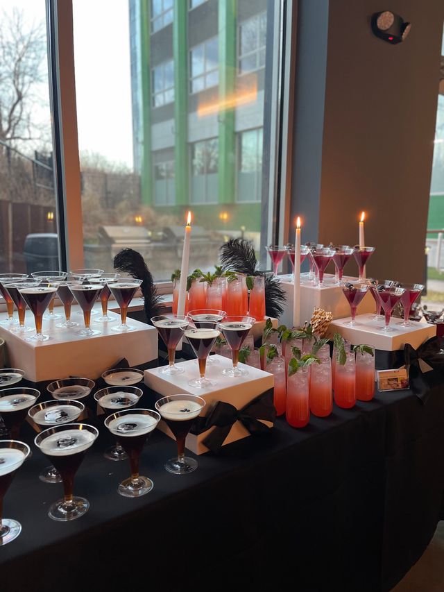 Stylish event cocktail station with rows of espresso-colored martini glasses and tall pink mint-garnished drinks on white risers, lit taper candles and black-feather accents by a large window with an urban building view.
