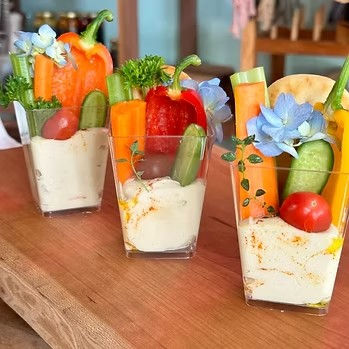Three clear cups of hummus-filled vegetable crudités with mini bell peppers, carrot sticks, baby cucumbers, cherry tomatoes, herbs and blue edible flowers arranged on a wooden board — colorful party appetizer.