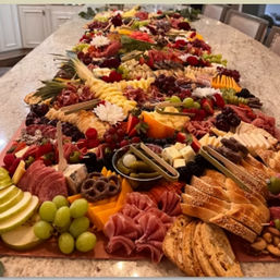 Long festive charcuterie grazing table on a kitchen island with assorted cheeses, cured meats, crackers, grapes, sliced fruit, pickles, nuts and floral garnishes — colorful party appetizer spread.