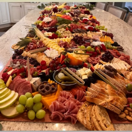 Long festive charcuterie grazing table on a kitchen island with assorted cheeses, cured meats, crackers, grapes, sliced fruit, pickles, nuts and floral garnishes — colorful party appetizer spread.