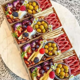 Four gourmet mini charcuterie boxes arranged diagonally on a marble board with green olives, rolled salami, crackers, cheeses, grapes, berries, and nuts