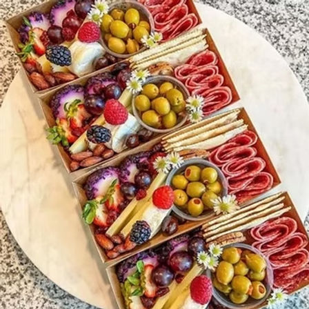 Four gourmet mini charcuterie boxes arranged diagonally on a marble board with green olives, rolled salami, crackers, cheeses, grapes, berries, and nuts