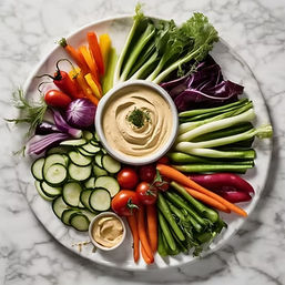 Colorful vegetable crudité platter with sliced cucumber, cherry tomatoes, carrots, bell peppers, radicchio, scallions and snap peas arranged around bowls of creamy hummus on a marble serving board