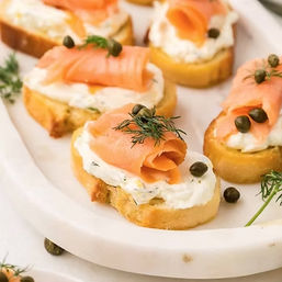 Smoked salmon crostini — toasted baguette rounds topped with herbed cream cheese, folded smoked salmon, capers and fresh dill on a white platter, perfect party or brunch appetizer.