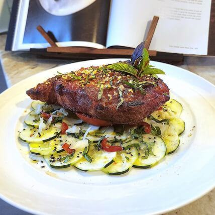 Golden crispy breaded cutlet on a bed of thinly sliced zucchini carpaccio with diced tomatoes, fresh herbs and basil garnish on a white plate, cookbook in background