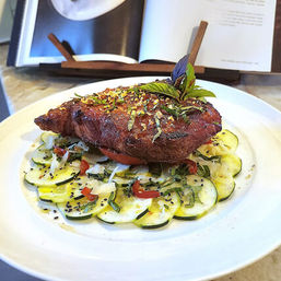 Golden crispy breaded cutlet on a bed of thinly sliced zucchini carpaccio with diced tomatoes, fresh herbs and basil garnish on a white plate, cookbook in background