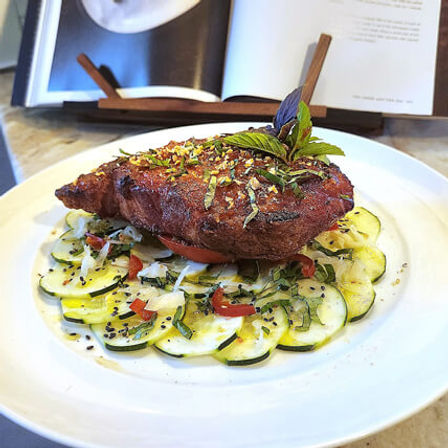 Golden crispy breaded cutlet on a bed of thinly sliced zucchini carpaccio with diced tomatoes, fresh herbs and basil garnish on a white plate, cookbook in background