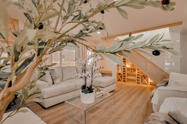 Airy modern cozy living room framed by leafy branches, featuring a neutral beige sectional, glass coffee table with potted orchids, light wood floors and built-in under-stair shelving and wine storage, softly lit by natural window light.