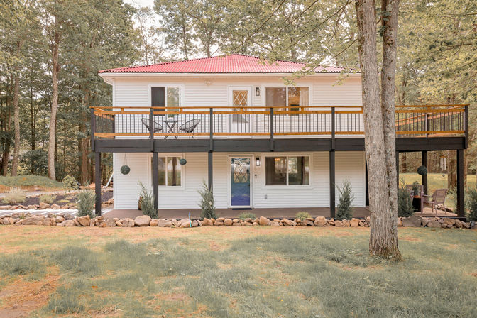 Charming two-story white country house with red metal roof, elevated wooden deck and blue front door, set on a grassy, tree-lined lot with stone-edged landscaping.
