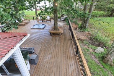 Spacious residential wooden backyard deck with built-in tree bench, inset hot tub, lounge chairs, fire pit and black metal railing overlooking a green wooded yard.