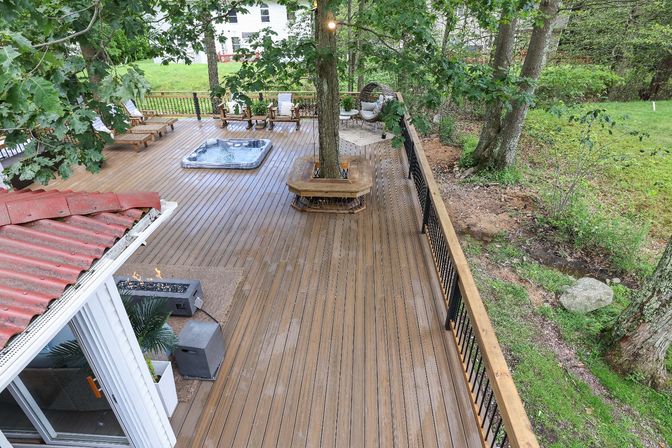 Spacious residential wooden backyard deck with built-in tree bench, inset hot tub, lounge chairs, fire pit and black metal railing overlooking a green wooded yard.