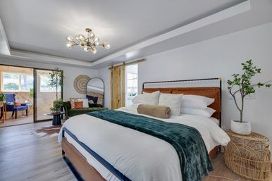 Bright modern master bedroom with leather headboard, crisp white bedding and teal throw, brass globe chandelier, sliding glass door to sunny sitting room, green velvet sofa, barn door and rattan side table with potted plant.