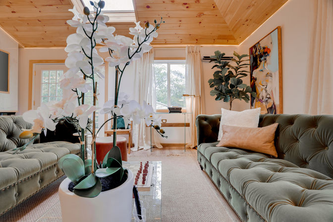 Bright cozy living room with wood-paneled vaulted ceiling and skylight, green tufted velvet sofas, white orchid centerpiece, fiddle-leaf fig and framed art.