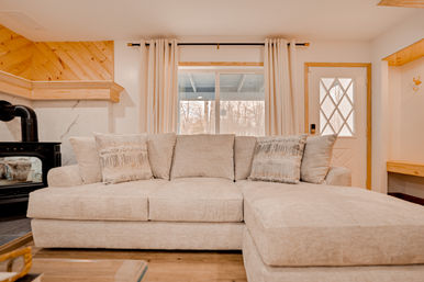 Cozy sunlit living room with large beige sectional sofa and patterned throw pillows, wood-burning stove, curtains framing a window overlooking backyard trees — modern rustic vibe