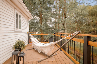 Cozy cream hammock with fringe on a wooden backyard deck beside a white house, overlooking a leafy forest with potted ferns and decorative lanterns.