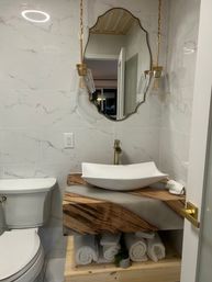 Modern small bathroom with marble tile walls, wood-and-resin floating vanity topped by a white vessel sink, gold faucet and pendant lights, decorative mirror and rolled white towels.