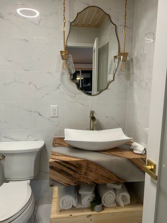Modern small bathroom with marble tile walls, wood-and-resin floating vanity topped by a white vessel sink, gold faucet and pendant lights, decorative mirror and rolled white towels.