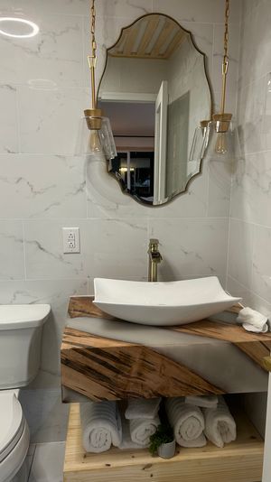 Modern small bathroom with marble tile walls, wood-and-resin floating vanity topped by a white vessel sink, gold faucet and pendant lights, decorative mirror and rolled white towels.