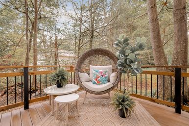 Cozy wooded backyard deck with a round rattan pod chair and tropical throw pillow, nested white side tables, potted plants, textured outdoor rug, and wooden railing overlooking trees