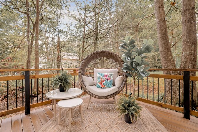 Cozy wooded backyard deck with a round rattan pod chair and tropical throw pillow, nested white side tables, potted plants, textured outdoor rug, and wooden railing overlooking trees