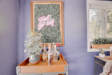 Home bar cart against a lavender wall with framed greenery and pink neon reading “Treat Yo Self,” bottles and cocktail shaker on a wooden tray, potted plant and nearby window planter