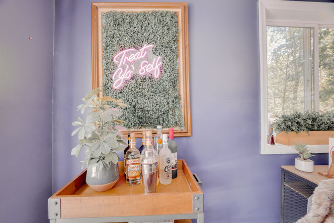 Home bar cart against a lavender wall with framed greenery and pink neon reading “Treat Yo Self,” bottles and cocktail shaker on a wooden tray, potted plant and nearby window planter
