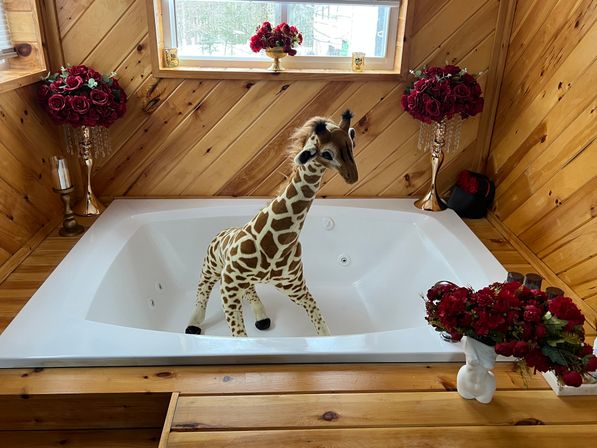 Stuffed giraffe toy standing in a white jetted bathtub inside a wood-paneled rustic cabin bathroom, surrounded by red rose bouquets and gold candleholders