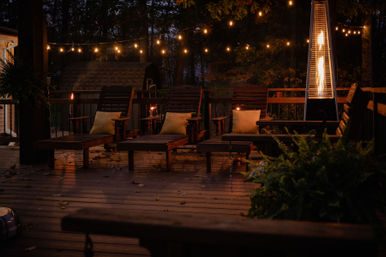 Cozy wooded backyard deck at dusk with wooden lounge chairs, glowing string lights, and a tall patio heater.
