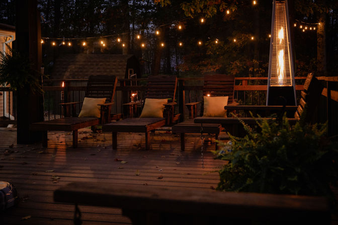 Cozy wooded backyard deck at dusk with wooden lounge chairs, glowing string lights, and a tall patio heater.