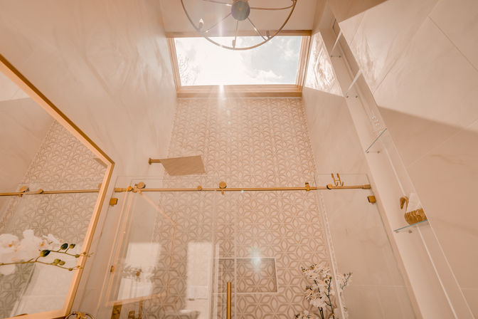 Sunlit luxury bathroom with skylight, glass-enclosed shower, geometric patterned wall tile, gold fixtures and white orchids for a bright, elegant feel.