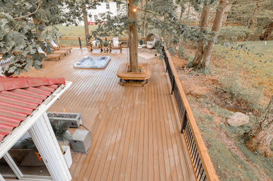 Aerial view of a spacious wooden backyard deck with a hot tub, lounge chairs, fire pit, wraparound tree bench and black metal railing, surrounded by trees and grassy yard.