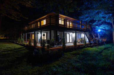 Night view of a cozy two-story rural house with a wraparound wooden deck and exterior staircase, warm interior lights and blue landscape uplighting illuminating patio furniture and surrounding trees.