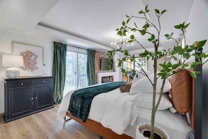 Cozy modern bedroom — leather headboard, plush white bedding with green velvet throw, potted indoor tree, sliding glass doors with green curtains, black accent cabinet, wood floors, wall-mounted TV and contemporary chandelier.