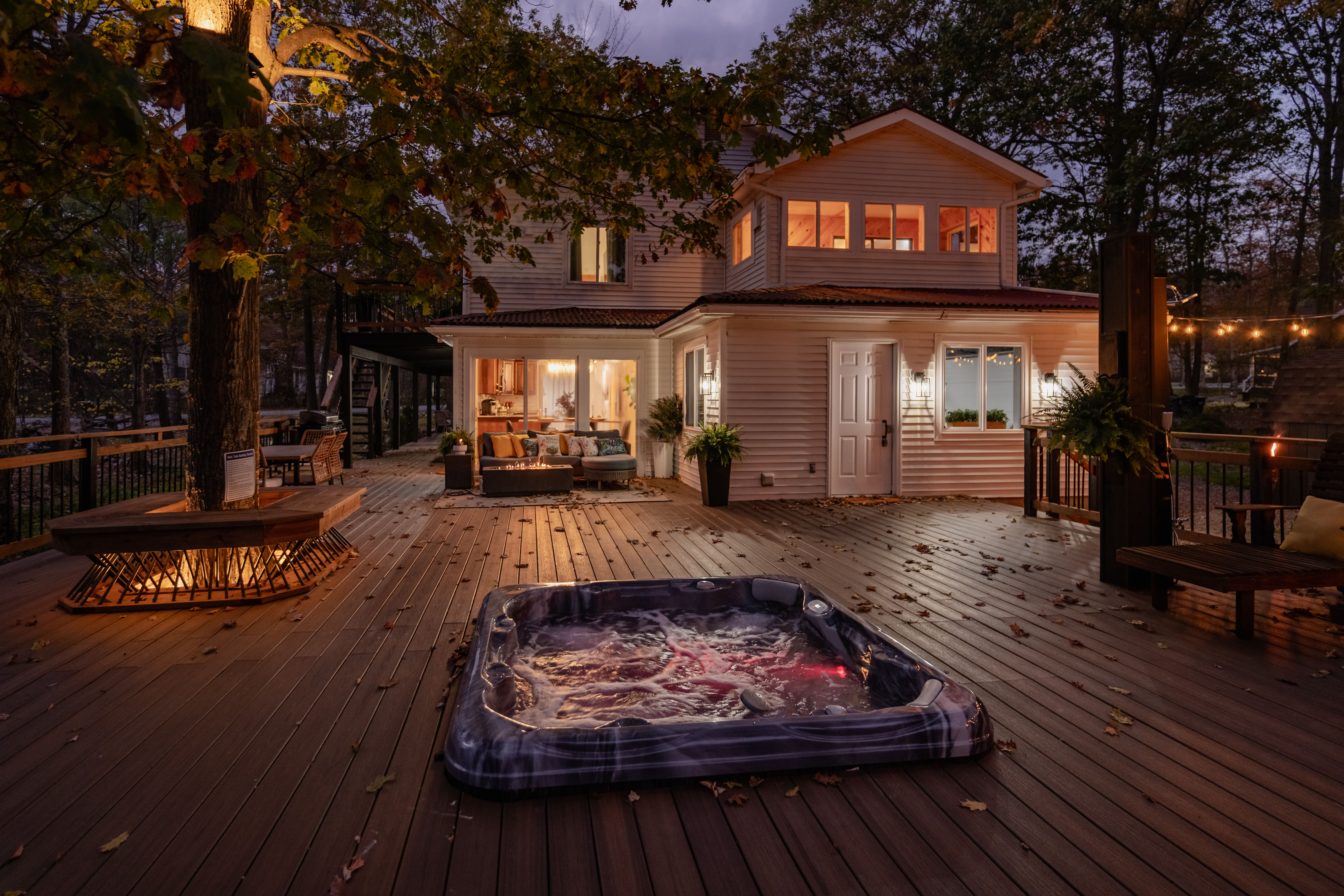 Evening backyard deck of a two-story suburban home in a wooded setting, bubbling hot tub on a wide wooden deck, cozy outdoor seating, fire pit, string lights and autumn leaves