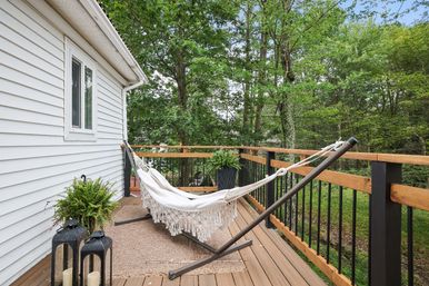 Cozy white fringed hammock on a metal stand on a wooden back deck beside white house siding, with potted ferns and lanterns, overlooking a lush wooded backyard