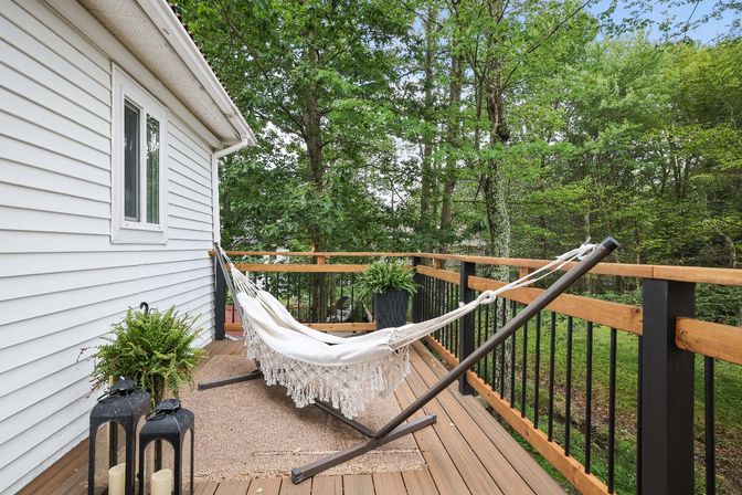 Cozy white fringed hammock on a metal stand on a wooden back deck beside white house siding, with potted ferns and lanterns, overlooking a lush wooded backyard