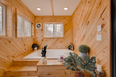 Cozy cabin-style wood-paneled bathroom with a sunlit drop-in soaking tub on a stepped wooden surround, windows framing bare winter trees, potted plants, candles and a rustic spa vibe.