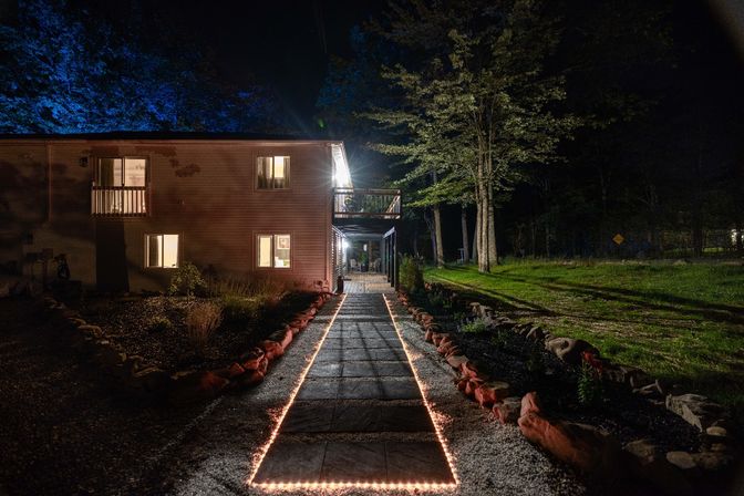 Warm LED-lined stone pathway leading to a two-story house with glowing windows and a covered deck, set in a wooded yard at night with blue uplighting on distant trees and stone borders.