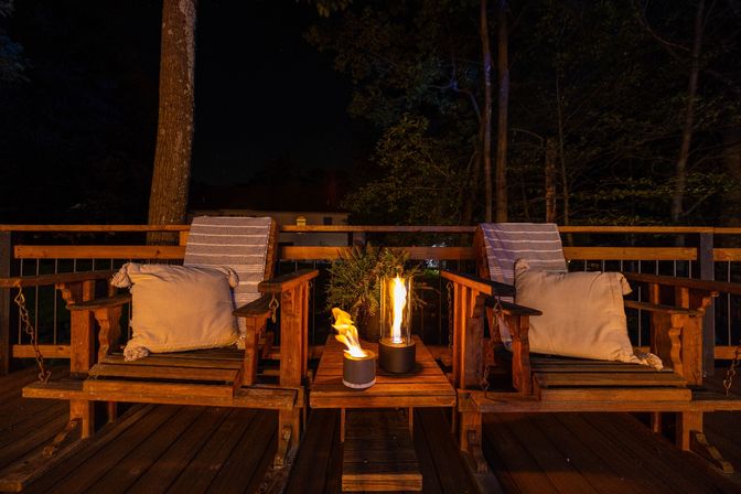 Cozy nighttime outdoor deck with two wooden lounge chairs, striped throws and cushions, and two small tabletop fire pits casting warm light
