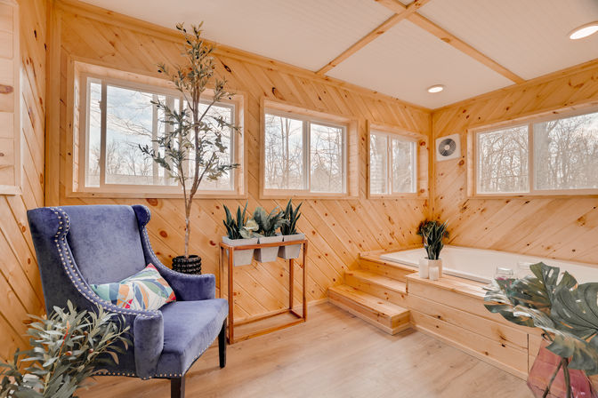 Bright wood-paneled relaxation room with a built-in soaking tub and steps, blue velvet wingback chair with a colorful pillow, potted plants, and large windows overlooking winter woods