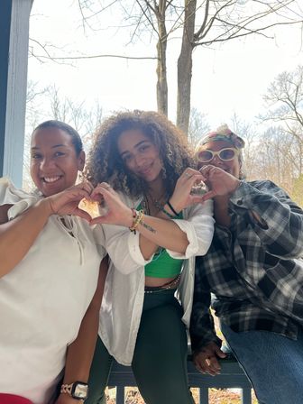Three friends sitting on a wooden porch in a wooded setting, smiling and making heart shapes with their hands — casual outdoor group photo.