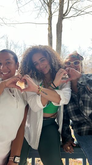 Three friends sitting on a wooden porch in a wooded setting, smiling and making heart shapes with their hands — casual outdoor group photo.